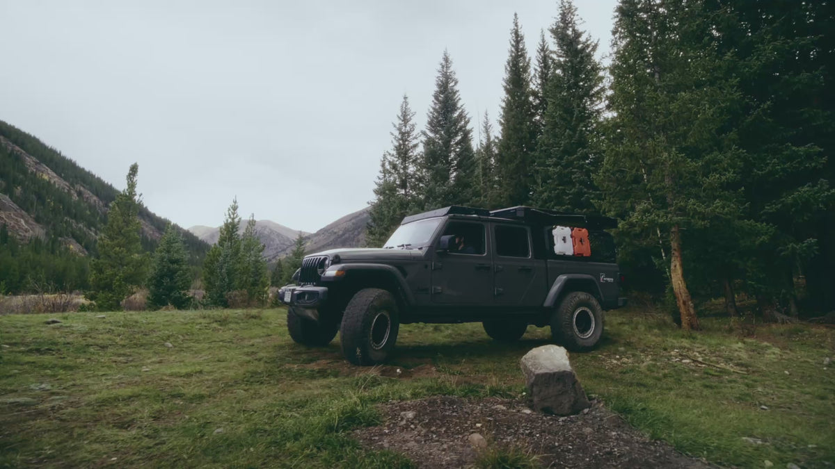 Load video: Truck tolling up in the mountains with a rooftop tent, with the sun setting. Two people are setting up camp for the evening, in an Intrepid Camp Gear tent, as the sun sets b